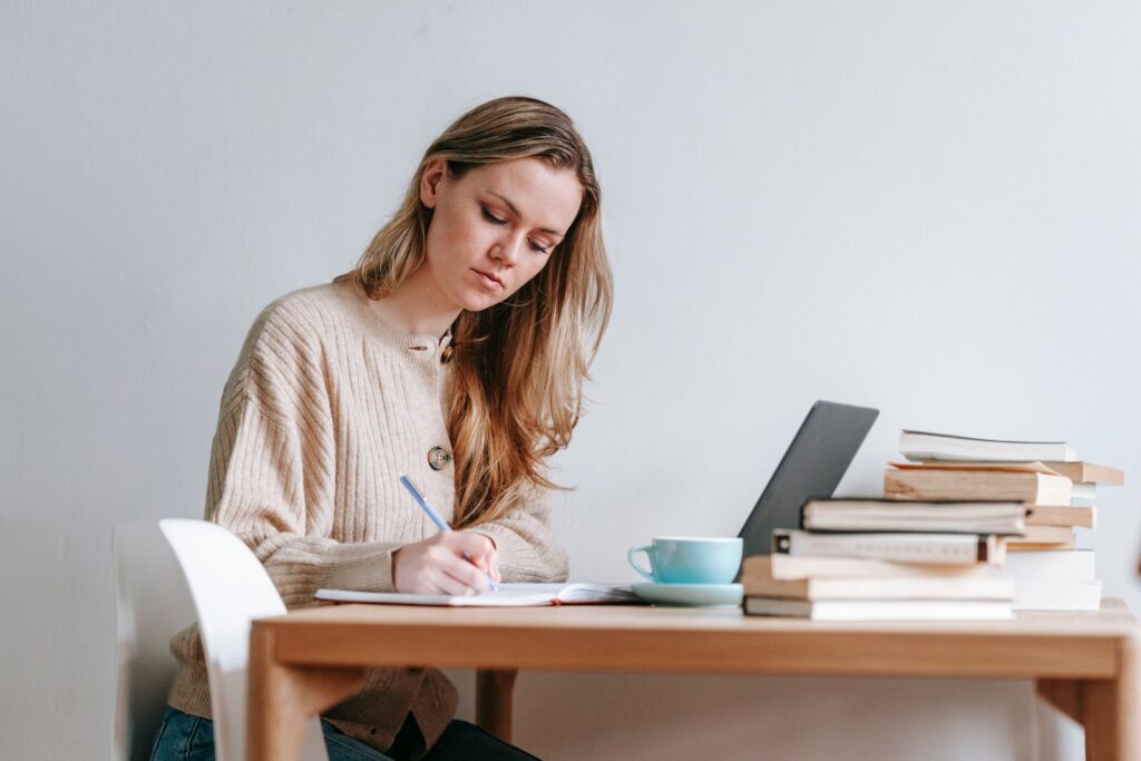 Woman writing in a notebook at a desk beside a laptop and a stack of books