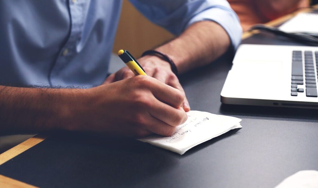 Hands writing notes on paper next to a laptop